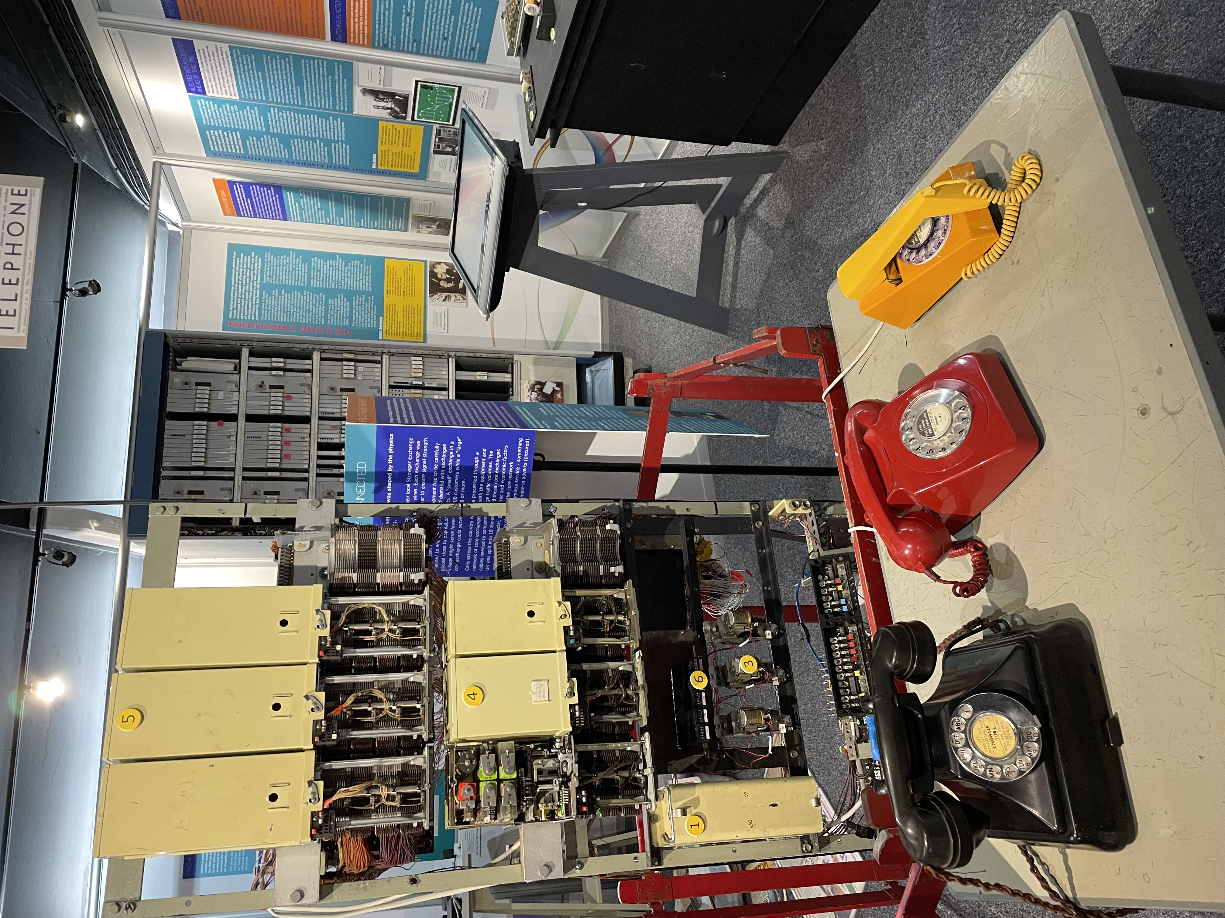 Interactive phone demonstration table with black, red and yellow telephones connected to exchange equipment